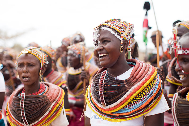Um grupo de mulheres Rendille em um festival. Todos estão vestidos com roupas tradicionais, elaboradas e coloridas com chapéus de miçangas. Cada um tem uma faixa listrada de cores vivas em volta dos ombros.