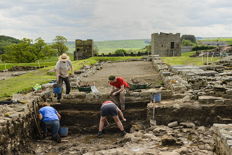 Quatre personnes creusent sur un site d'excavation. Ils ont partiellement découvert les fondations et le sol en pierre d'un ancien bâtiment. Deux structures carrées en pierre sont visibles en arrière-plan.
