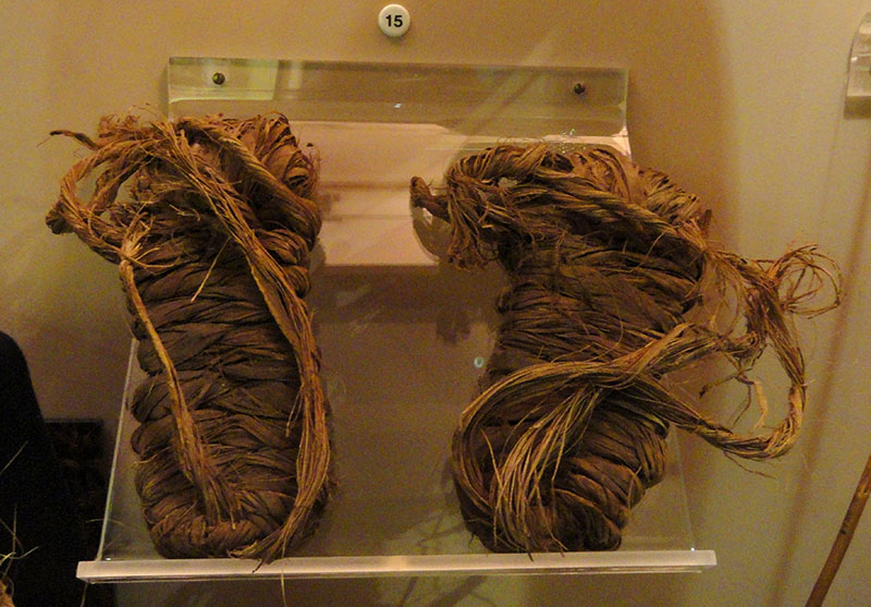 Une paire de sandales exposée derrière une vitre. Les sandales sont fabriquées en matière végétale torsadée et tressée.