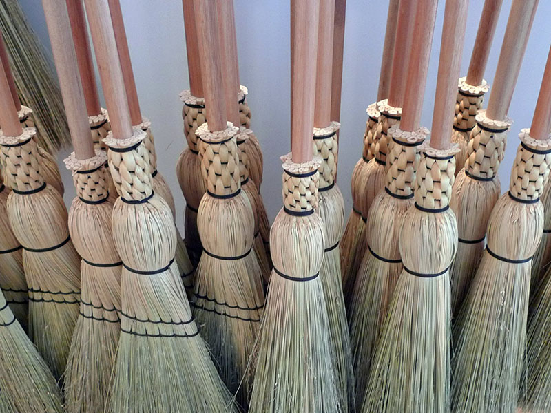 Uma fotografia colorida de um grupo de cerca de quinze vassouras de palha. As vassouras têm alças iguais de madeira lisa. O fundo é construído com uma fibra vegetal de cor clara. As fibras são presas às vassouras usando fio escuro.
