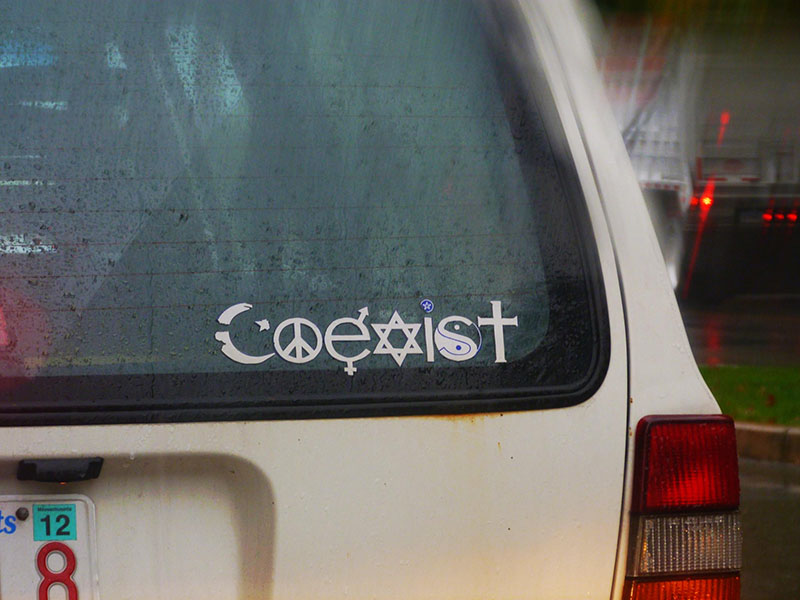 Photographie en couleur d'un autocollant de pare-chocs à l'arrière d'une voiture, épelant le mot « Coexister » à l'aide de divers symboles. La lettre « c » est remplacée par un croissant de lune représentant l'islam. La lettre « o » est remplacée par le symbole de la paix. Pour la lettre « e », un symbole masculin/féminin est utilisé. Pour la lettre « x », apparaît l'étoile de David représentant le judaïsme. Pour la lettre « i », un symbole païen/WICCAN est utilisé. La lettre « s » est remplacée par un symbole chinois yin-yang. Pour la lettre « t », une croix représentant le christianisme est utilisée.