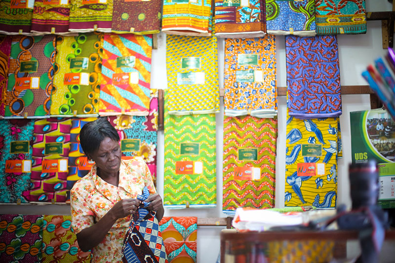 Photographie en couleur d'une femme debout devant un mur présentant de longs tissus colorés ornés de grands motifs aux couleurs vives. La femme tient du tissu qu'elle semble coudre à la main.