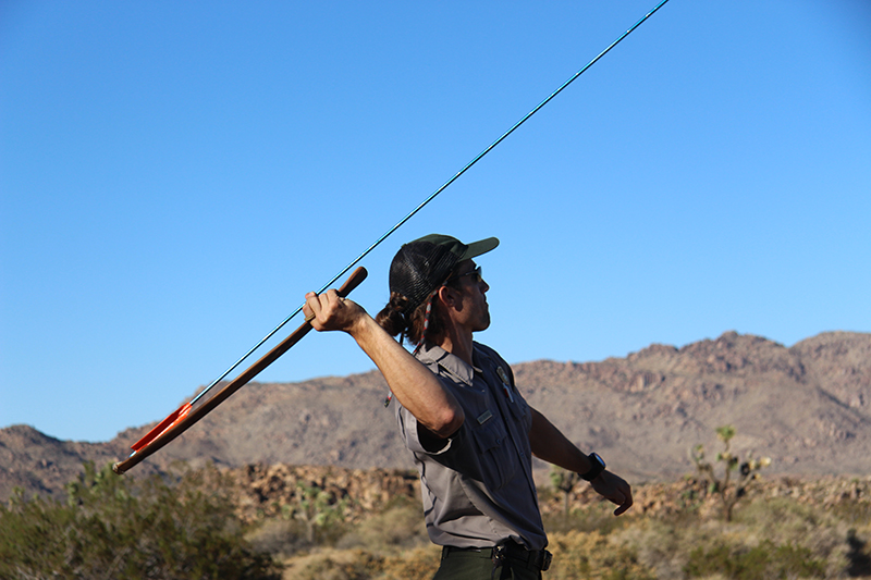 A man holds the atlatl behind his back, with a long spear notched into the edge furthest from his hand. He appears poised to launch the spear.