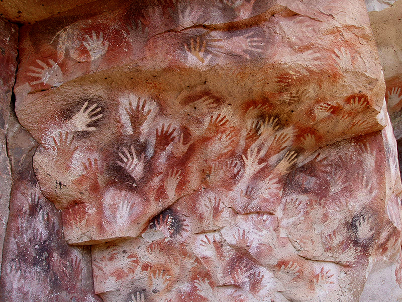 Parede de caverna decorada com dezenas de impressões de mãos sobrepostas em várias cores e tonalidades.