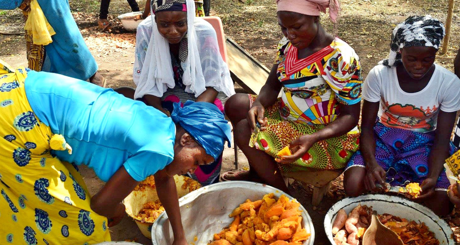 Quatro mulheres africanas vestindo roupas tradicionais sentadas em círculo do lado de fora, atrás de duas grandes tigelas de batata-doce cozida.