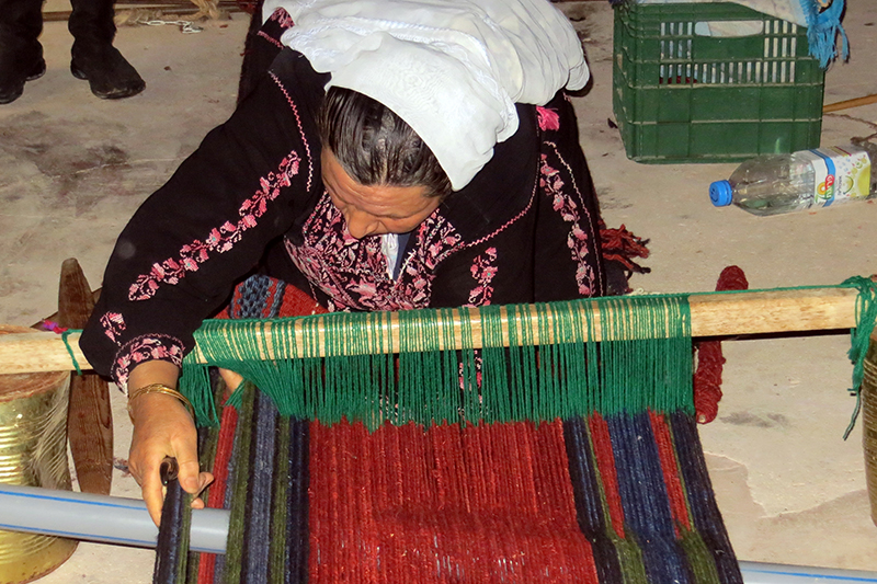 Una mujer con un pañuelo en la cabeza sentada detrás de una simple máquina de madera tejiendo lana.