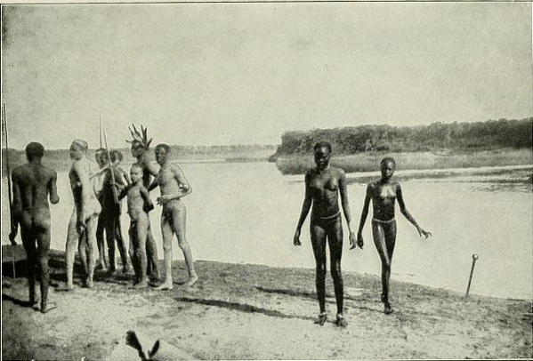 Uma foto em preto e branco de membros do povo Nuer tirada em 1906. Eles estão juntos na margem de um rio, a maioria em um aglomerado, mas com um par de pares sozinhos.