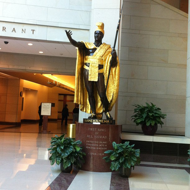 Uma estátua de um homem em pé com a mão estendida em um gesto oratório. Ele segura um bastão na outra mão. A estátua está vestindo uma capa e um chapéu dourados e fica em um espaço interno de aparência contemporânea.