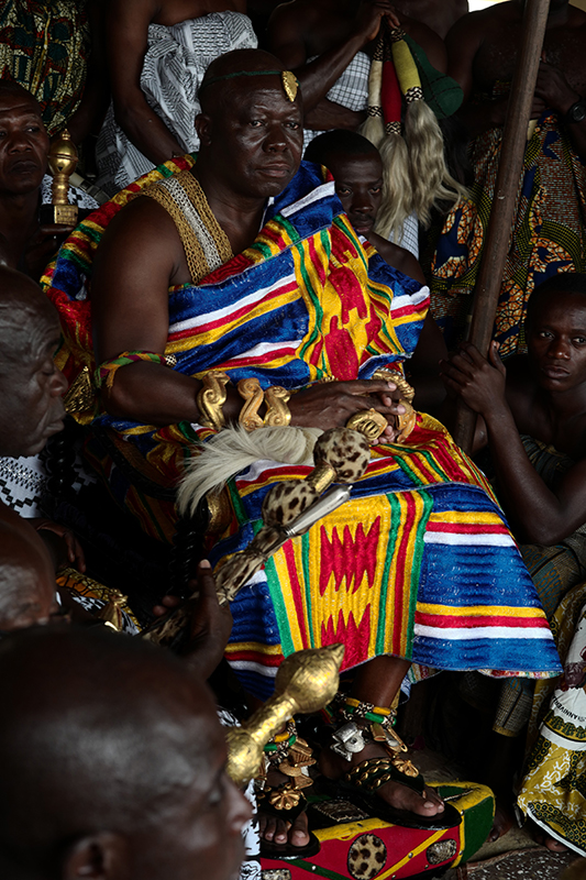 رجل أفريقي جالس، يرتدي رداءً بألوان زاهية ومنقوش بجرأة وأساور ذهبية سميكة. تعبيره مدروس وجاد.