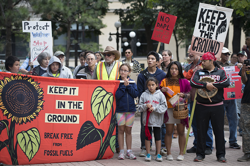 Um grupo de pessoas, incluindo muitas crianças, se envolveu em uma marcha de protesto. Um banner proeminente diz “Mantenha-o no chão - liberte-se dos combustíveis fósseis”.