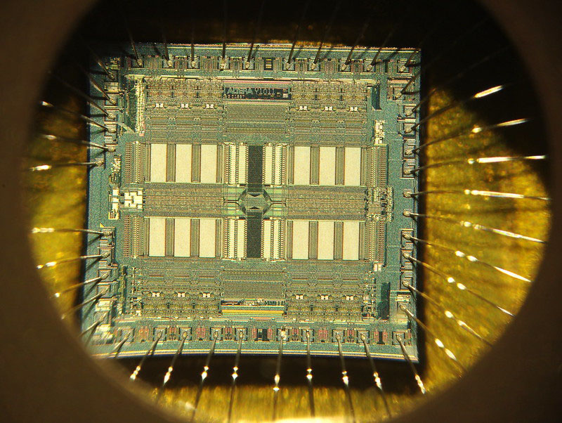 Magnified color photograph of a semiconductor chip. The magnification makes visible a complex pattern of shapes and lines across the surface of the chip.