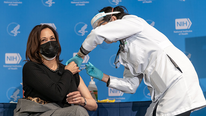 Color photograph of Kamala Harris holding up the sleeve of her shirt to bare her shoulder while a woman in a white lab coat administers a shot. In the background is a board displaying the logo of the National Institutes of Health.