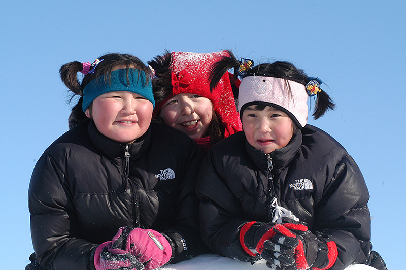 Três jovens inuítes vestidas com casaco de inverno, luvas e faixas de ouvido estão sentadas juntas e sorrindo.