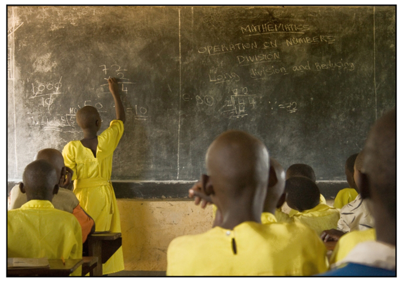 Uma garota masaii vestindo um uniforme escolar amarelo está resolvendo um problema de matemática no quadro-negro, enquanto os outros alunos estão sentados na sala de aula.