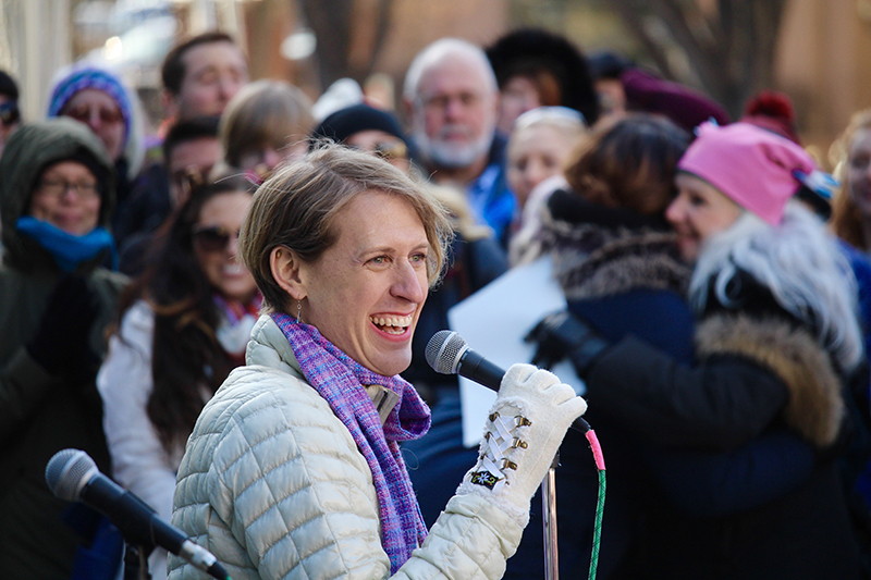 Une femme vêtue de vêtements d'hiver debout tenant un microphone lors d'un événement en plein air. Une foule toute vêtue de vêtements d'hiver se tient derrière elle.