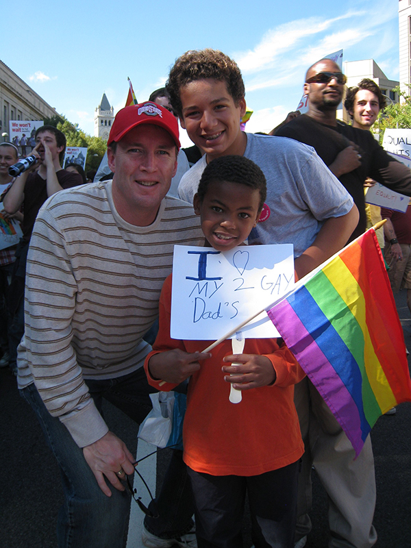 一个白人、一个十几岁的男孩和一个年轻的黑人男孩正在聚会上摆姿势拍照。 那个黑人男孩拿着一个标语，上面写着 “我爱我的两个爸爸”。 他还拿着彩虹旗。