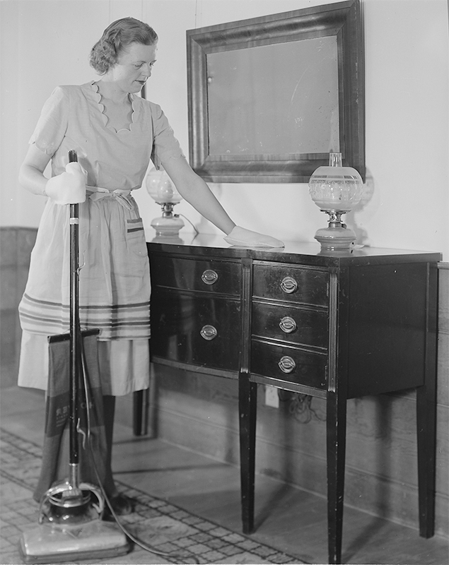 Uma mulher americana está limpando o pó de um aparador. Ela está segurando um aspirador elétrico na outra mão.