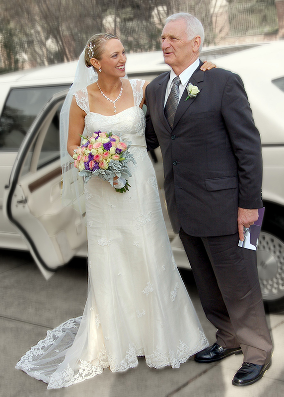 عروس ترتدي فستان زفاف أبيض طويل، تقف بجانب رجل ناضج يرتدي بدلة قطنية. كلاهما يقفان أمام سيارة ليموزين بيضاء.
