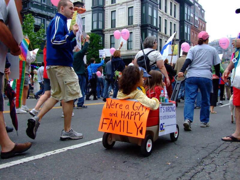 A group of people walking in a parade. A woman is pulling a wagon with several children inside. A sign on the back reads We are a gay and happy family.