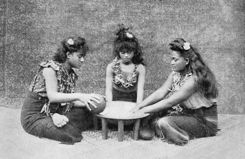 Três jovens samoanas vestindo roupas tradicionais estão sentadas em uma pequena mesa jogando um jogo.