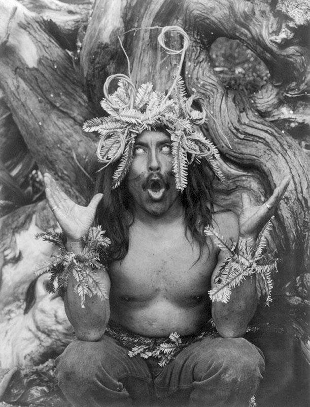 Black and whte image of a man with his eyes rolled back so that only the whites are visible. He holds his hands at shoulder height, palms up. He wears a headdress and bracelets made of plant material. Behind him is visible the twisted trunk of a tree.