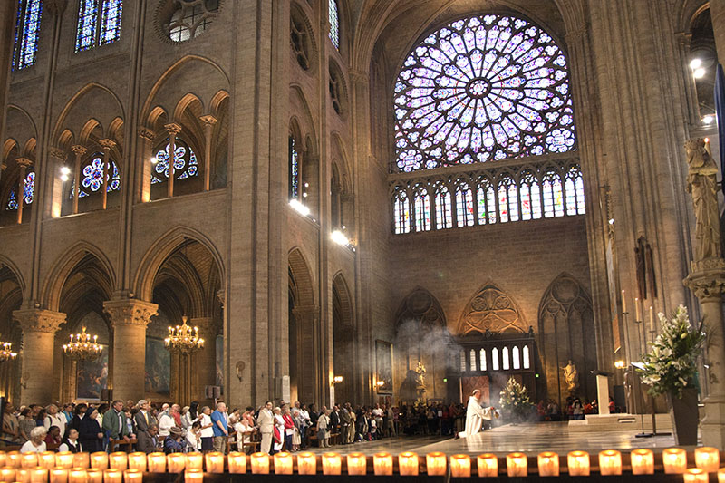 Imagem interior da catedral de Notre Dame durante um serviço religioso. Dois níveis de arcos góticos são visíveis e acima deles estão os vitrais em forma de medalhões. As figuras humanas na imagem são muito pequenas, enfatizando o tamanho da catedral.