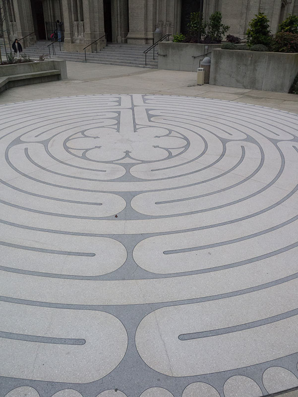 Un motif intégré dans un sol en pierre. Le motif est créé par des lignes claires sur une surface plus foncée. Les lignes tracent des sentiers de randonnée dans un cercle. Les chemins mènent finalement à une forme de fleur au centre du cercle.
