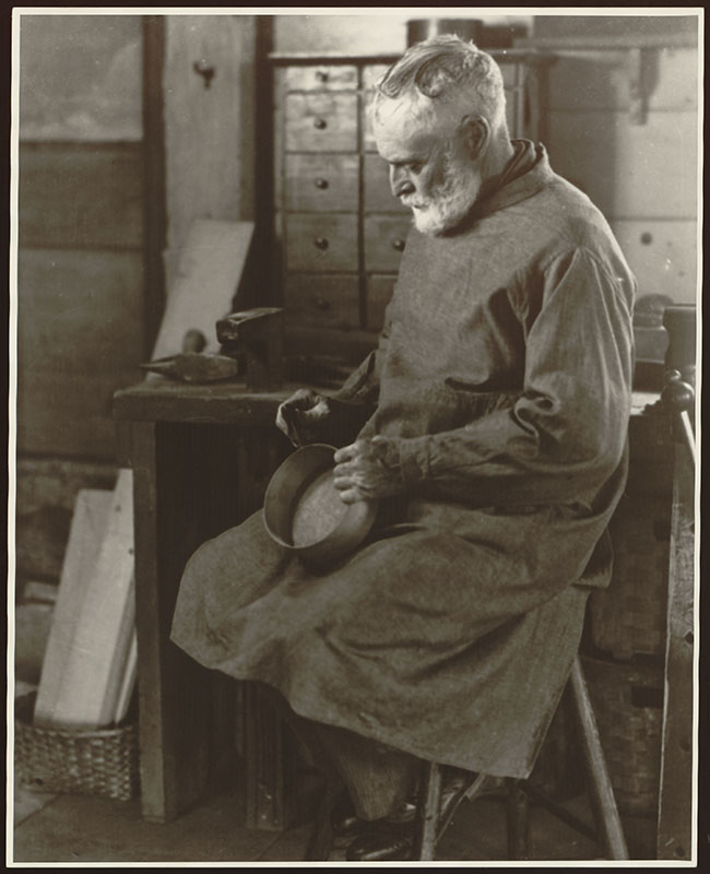 Un hombre se sienta en un banco sosteniendo una caja ovalada en sus manos. Viste una bata hasta la rodilla. Las herramientas para trabajar la madera son visibles en la mesa detrás de él.