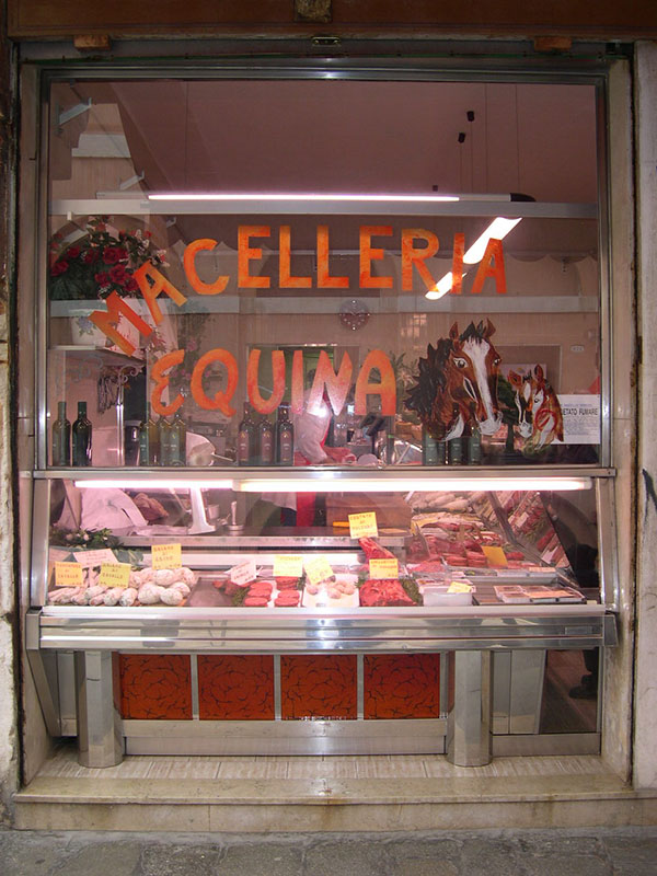 نافذة متجر الجزار مع قطع اللحم مرئية في علبة عرض خلف الزجاج. تقول اللافتة الموجودة على النافذة، ما سيليريا إكوينا.