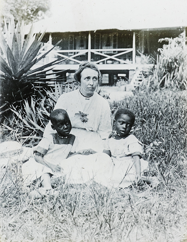 Fotografia em preto e branco de uma mulher branca sentada na grama em frente a uma casa, com duas crianças negras sentadas de cada lado. Todos os três usam expressões solenes. Um edifício simples com estrutura de madeira é visível ao fundo.