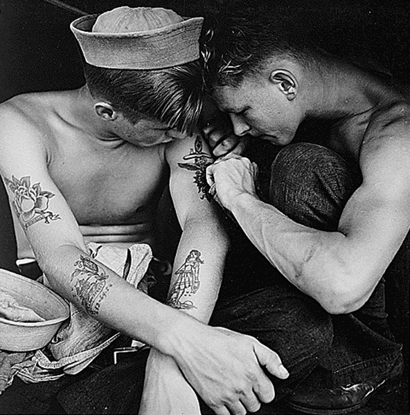 Imagem em preto e branco de dois jovens marinheiros, um tatuando o braço do outro.