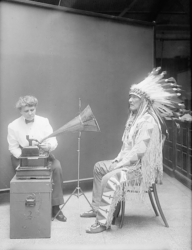 一个穿着正装外套和裤子的女士坐在留声机后面。 一个戴着全头饰的美洲原住民男子坐在留声机的扬声器前。