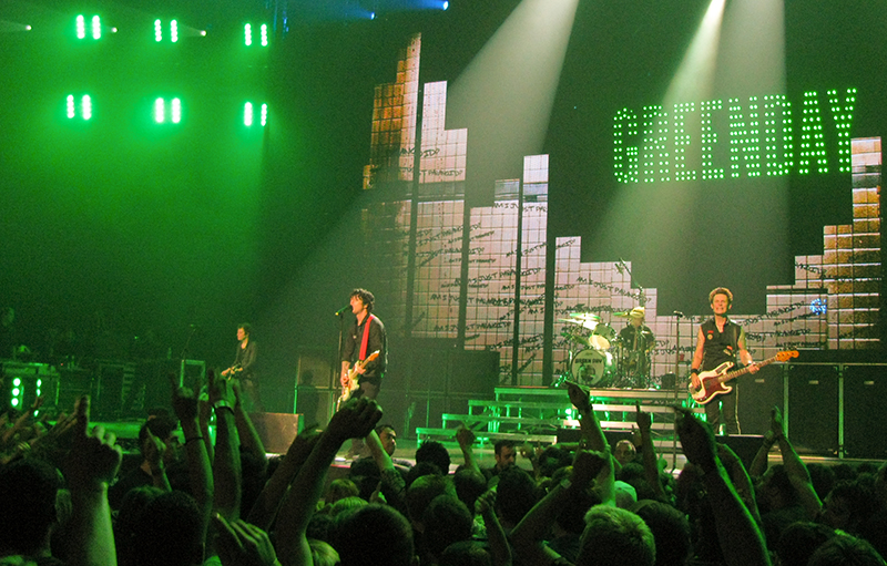 Banda se apresentando em um grande palco com uma multidão animada em primeiro plano.