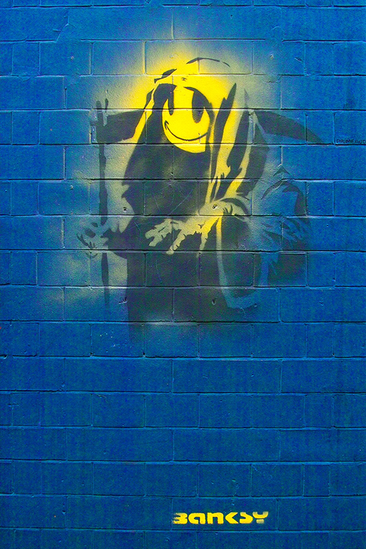 Imagem pintada com spray na parede de tijolos da figura Grim Reaper com um rosto sorridente amarelo brilhante.