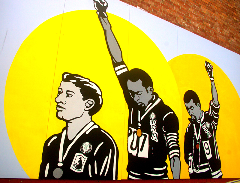 墙上的壁画，描绘了三名运动员脖子上挂着奖牌。 两个人低下头，举起拳头向空中。