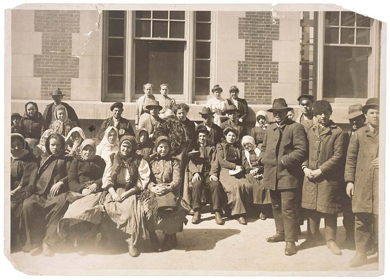 Photographie en couleur sépia d'un groupe de personnes européennes à l'extérieur d'un bâtiment à Ellis Island. Ils sont habillés simplement et semblent attendre.