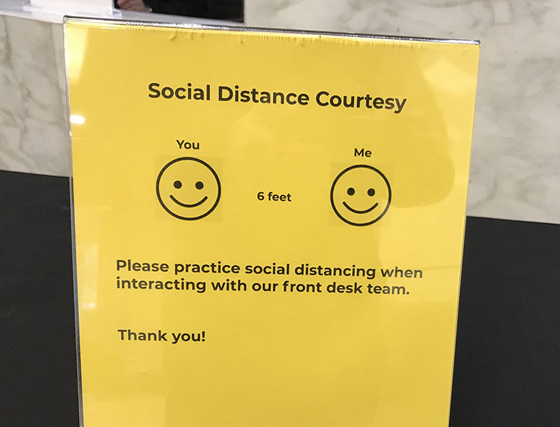Une affiche intitulée Social Distance Courtesy. Sous le titre apparaissent deux visages souriants, l'un étiqueté « Toi » et l'autre « Moi ». Dans l'espace entre les faces est imprimé la mention « 6 pieds ». Le texte au bas du dépliant se lit comme suit : « Veuillez respecter la distanciation sociale lorsque vous interagissez avec notre équipe de réception. Merci ! »
