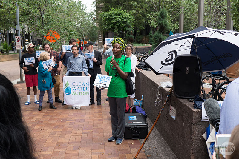 Un groupe de personnes qui organise un rassemblement dans un espace public. Ils brandissent des pancartes sur lesquelles on peut lire « Clean Air Now » et « Clean Water Now ». Une femme tient un micro et parle.