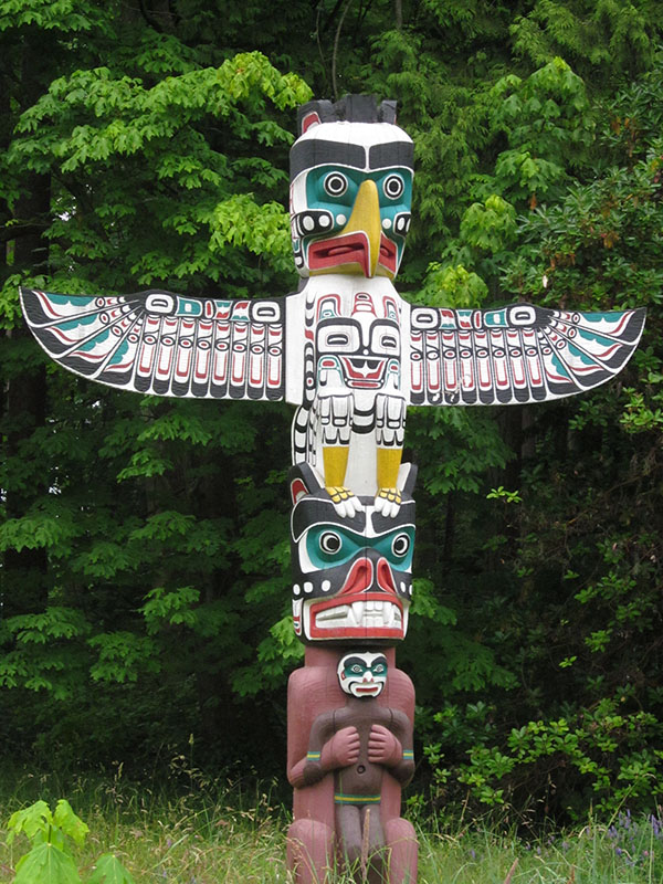Mât totémique avec deux personnages empilés l'un sur l'autre. La figure supérieure ressemble à un oiseau, avec de grandes ailes déployées. La figurine du bas tient une silhouette plus petite contre sa poitrine.