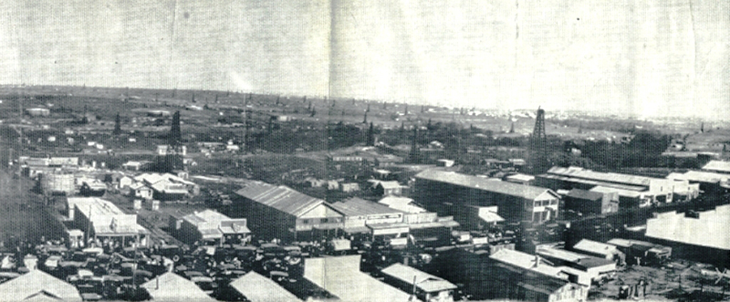 Vista aérea de muitos edifícios longos ao longo de uma rua principal. Aproximadamente duas dúzias de torres de petróleo são visíveis ao fundo.