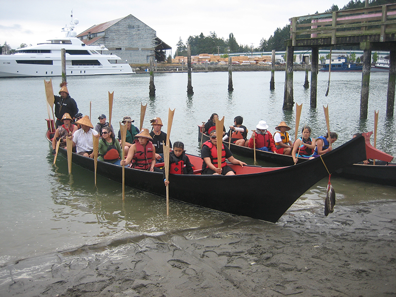 Duas canoas cheias de pessoas situadas perto de uma praia. As pessoas seguram os remos na posição vertical, usando as pontas para empurrar contra o fundo do lago.