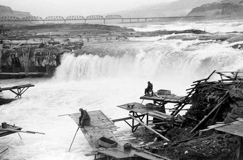 Deux hommes sont assis sur des plateformes en bois au bord d'une rivière, tenant de longues perches dont les extrémités sont immergées dans l'eau. Une cascade est visible derrière eux.