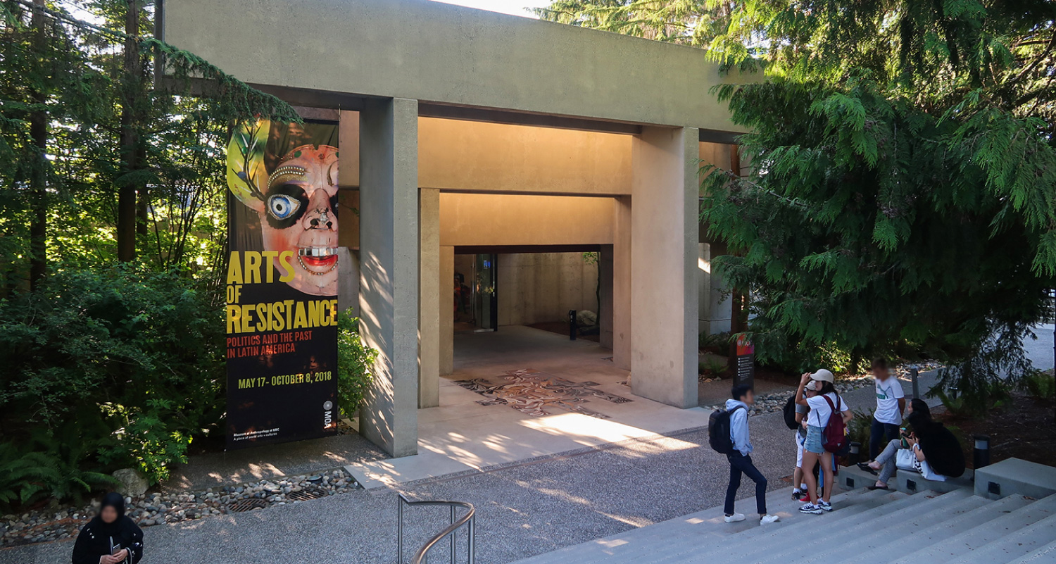 不列颠哥伦比亚省人类学博物馆的正门。 开口是一个大矩形，由通往门的渐变尺寸的矩形水泥柱勾勒出来。 设计现代。 左边的标语宣传一个名为 “抵抗艺术” 的展览。 几个人聚集在大楼前的楼梯上。
