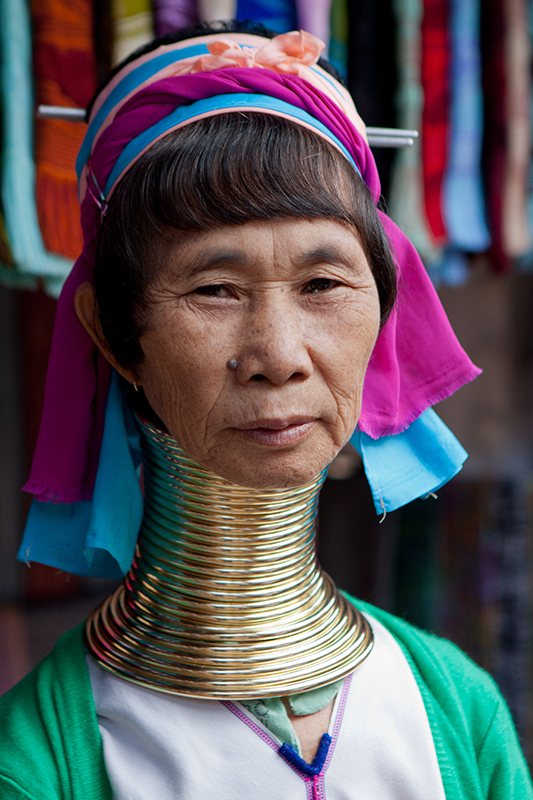 Woman with numerous gold neck rings stacked from her shoulders to underneath her jaw. There are about 23 rings. They are larger at her shoulders and become smaller as they move toward her jaw. The rings have lengthened her neck noticeably.