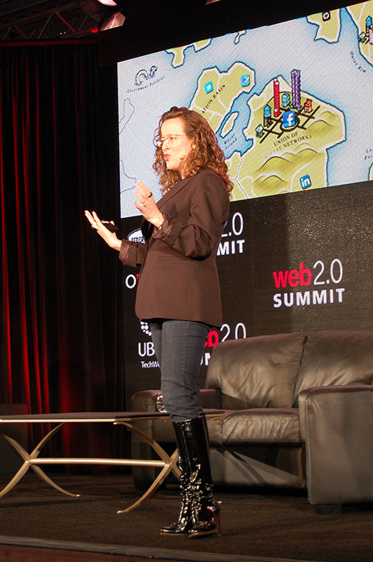 Uma mulher com longos cabelos cacheados e vestindo um blazer, jeans e botas está em um palco falando em uma conferência.