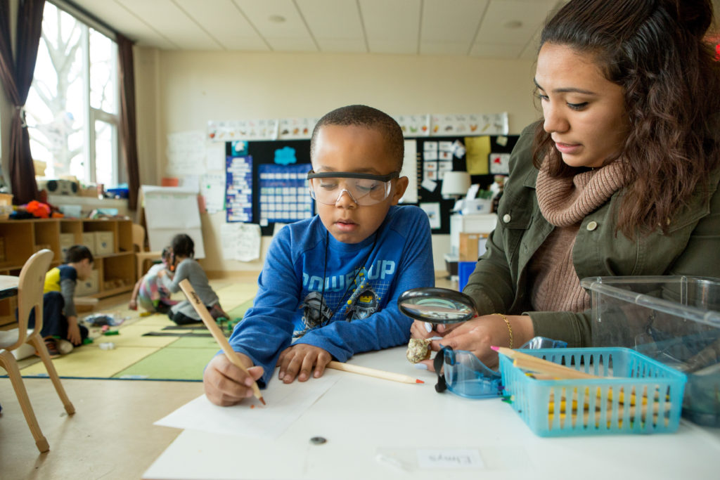 Un joven con gafas protectoras escribe con un lápiz en una hoja de papel mientras su maestro sentado a su lado sostiene una lupa.