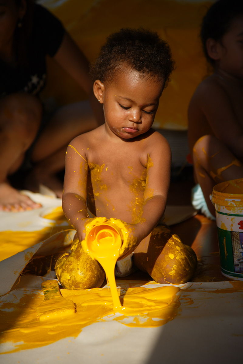 An infant kneels on a large piece of paper and pours yellow paint onto it. 