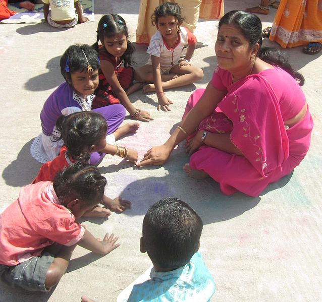 A woman sits on the ground with several children around her, laying their fingers on the ground. 