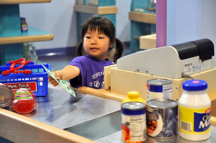 A young girl plays pretend at a dramatic play grocery store, handing play money over the counter. 