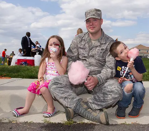 Un militar se sienta en una acera con dos niños comiendo bocadillos.